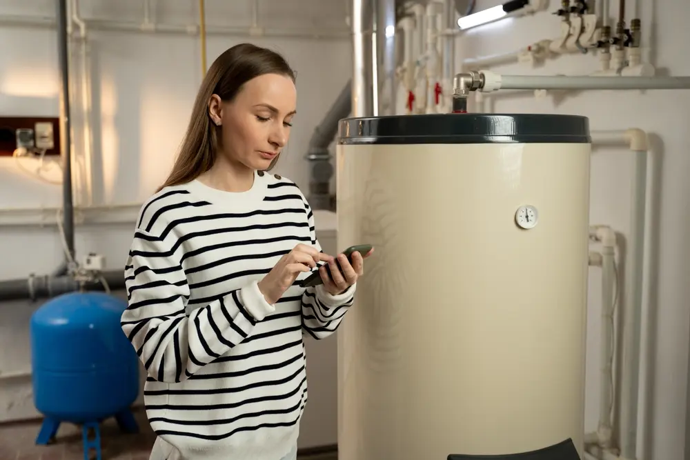 A young woman examines a non-operational boiler with concern, holding a phone in her hand, likely seeking help or planning to call for repair services.A young woman examines a non-operational boiler with concern, holding a phone in her hand, likely seeking help or planning to call for repair services.